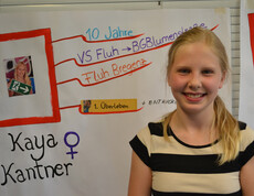 Eine Jugendliche mit blonden langen Haaren und gestreiften Oberteil steht vor einem Flipchart. Sie lächelt in die Kamera. 