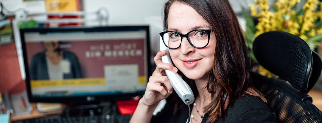 Eine junge Frau sitzt vor einem Computer und hält einen Telefonhörer in der Hand.
