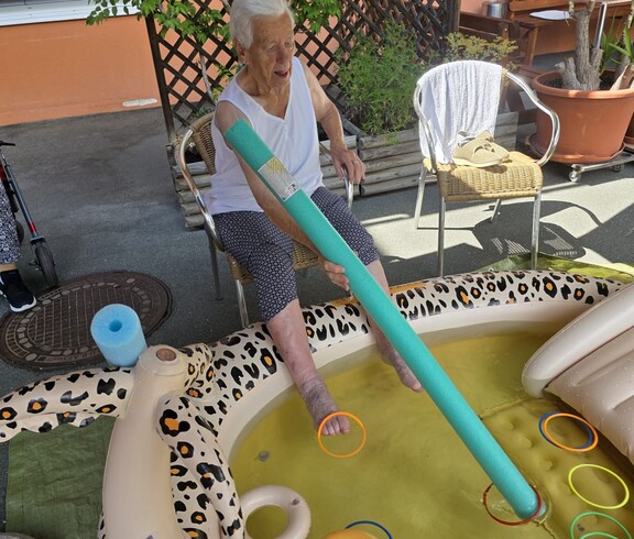 Auf der Terrasse des Pflegewohnhauses sitzt eine Bewohnerin vor einem aufblasbaren Planschbecken und hält eine lange Schaumstoffstange, während beide Füße im Wasser eingetaucht sind. Im Becken schwimmen bunte Ringe und andere Wasserutensilien, mit denen ein spielerisches Geschicklichkeitstraining durchgeführt wird