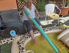 Auf der Terrasse des Pflegewohnhauses sitzt eine Bewohnerin vor einem aufblasbaren Planschbecken und hält eine lange Schaumstoffstange, während beide Füße im Wasser eingetaucht sind. Im Becken schwimmen bunte Ringe und andere Wasserutensilien, mit denen ein spielerisches Geschicklichkeitstraining durchgeführt wird