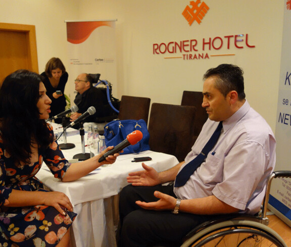 Ein Journalist interviewt einen Mann bei einer Veranstaltung von Caritas Albanien im Rogner Hotel Tirana, im Hintergrund ein Mann im Rollstuhl.