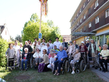 Bewohnerinnen und Bewohner eines Pflegewohnhauses versammeln sich gemeinsam mit Mitarbeitenden rund um den festlich geschmückten Maibaum und genießen das traditionelle Beisammensein im Freien.