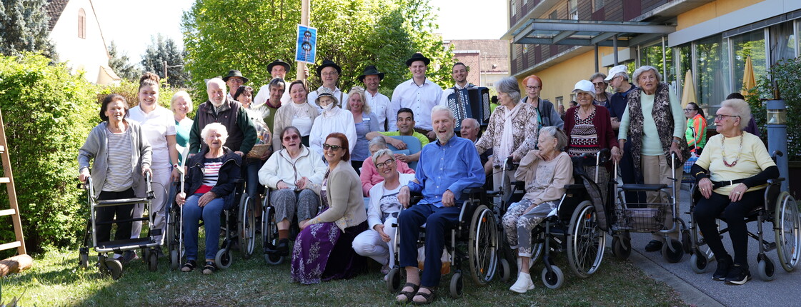 Bewohnerinnen und Bewohner eines Pflegewohnhauses versammeln sich gemeinsam mit Mitarbeitenden rund um den festlich geschmückten Maibaum und genießen das traditionelle Beisammensein im Freien.