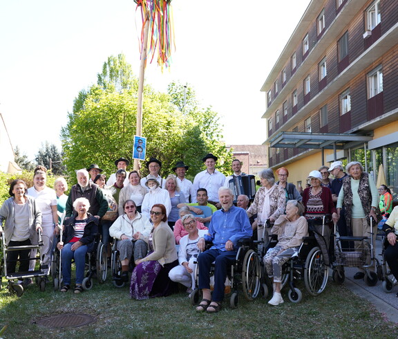Bewohnerinnen und Bewohner eines Pflegewohnhauses versammeln sich gemeinsam mit Mitarbeitenden rund um den festlich geschmückten Maibaum und genießen das traditionelle Beisammensein im Freien.