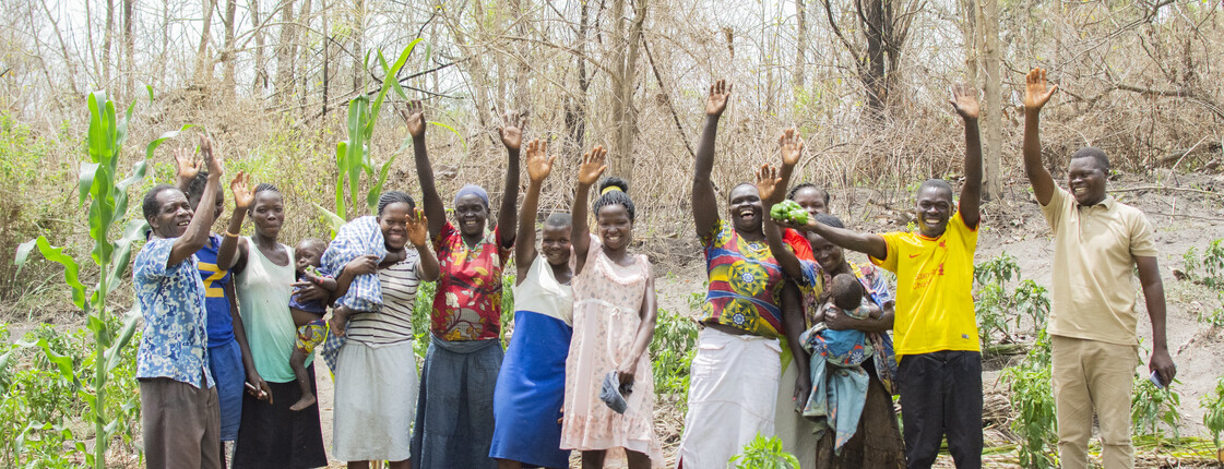 Mehrere Erwachsene aus dem Südsudan stehen lächelnd auf einem Feld vor Bäumen und heben lächelnd ihre Arme in die Luft.