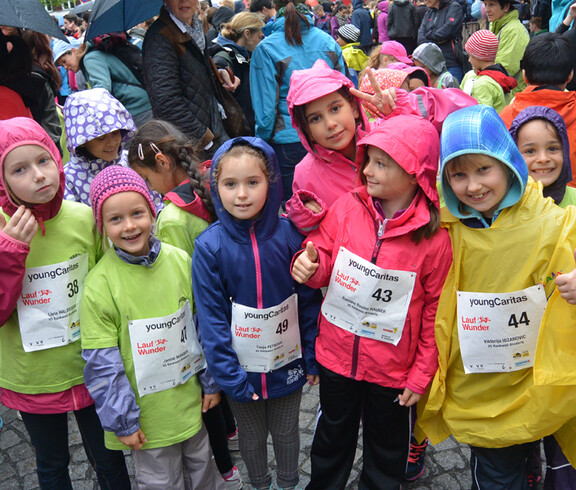 Etliche kleine Kinder nehmen bei einem Laufwettbewerb in der feldkircher Innenstadt teil. Sie tragen alle Startnummern, die jeweils an der Brust befestigt wurden, und unterschliedlich farbige Regenjacken und Trikots. 