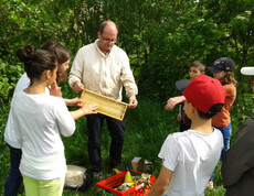 Sechs Kinder im Volksschulalter stehen um einen erwachsenen Mann. Sie sind im Freien bei einem Wald. Der Herr ist Imker und zeigt den Kindern eine Wabe. Die Kinder sind gespannt und sehr aufmerksam. Sie sind sommerlich bekleidet. 