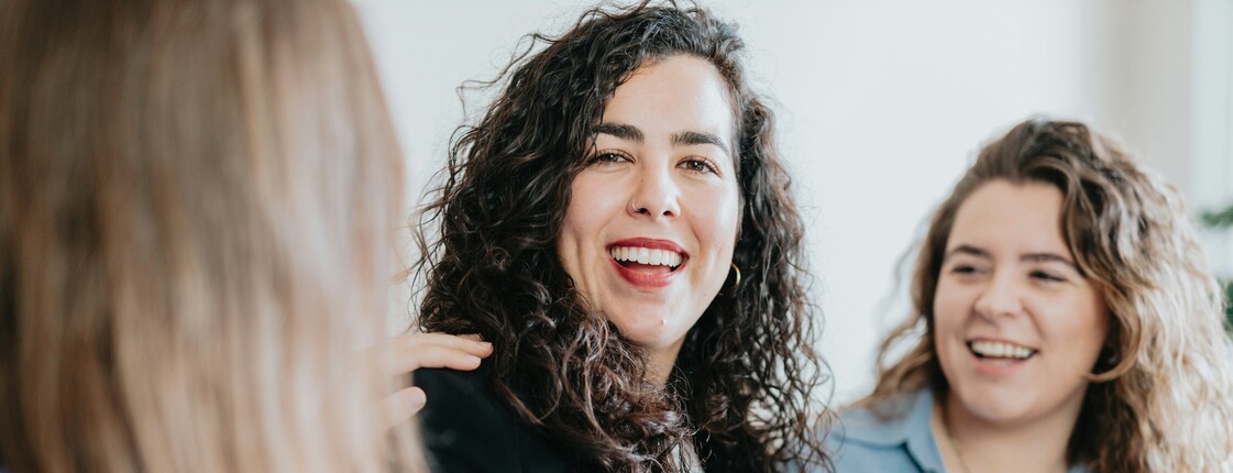 Zwei junge Frauen lachen in die Kamera, während im Vordergrund die Rückenansicht einer weiteren Frau zu sehen ist.