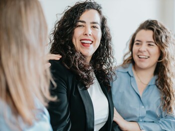 Zwei junge Frauen lachen in die Kamera, während im Vordergrund die Rückenansicht einer weiteren Frau zu sehen ist.
