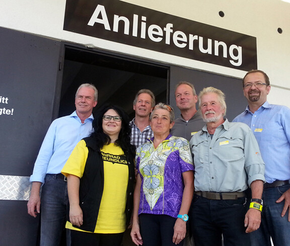 carla Mitarbeiter stehen zu einem Gruppenfoto zusammen. Sie sind beim Bereich der Warenanlieferung zusammengestanden. Sowohl die Männer als auch die Frauen sind gemütlich angenehm gekleidet.  