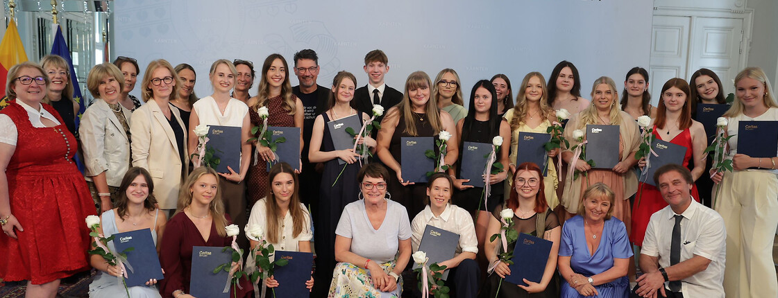 Große Gruppe von Absolvent*innen mit Urkunden und weißen Rosen vor Wand mit „Land Kärnten“