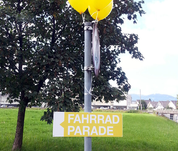 Ein gelb-weißes Schild auf einem metallernem Pfosten wurde angebracht. Darauf zu lesen: Fahrradparade. Auch hängen am Pfosten gelbe Luftballons. 