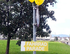 Ein gelb-weißes Schild auf einem metallernem Pfosten wurde angebracht. Darauf zu lesen: Fahrradparade. Auch hängen am Pfosten gelbe Luftballons. 