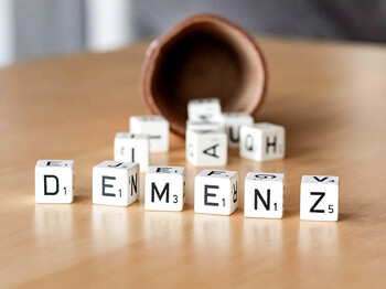 Eine brauner Becher liegt umgekippt auf einem Holztisch. Daneben liegen Würfel mit Buchstaben. Sie bilden das Wort „DEMENZ“.