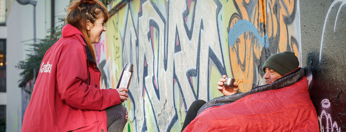 Eine Caritas-Mitarbeiterin bringt einem obdachlosen Mann warmen Tee – Hilfe und Menschlichkeit direkt auf der Straße.