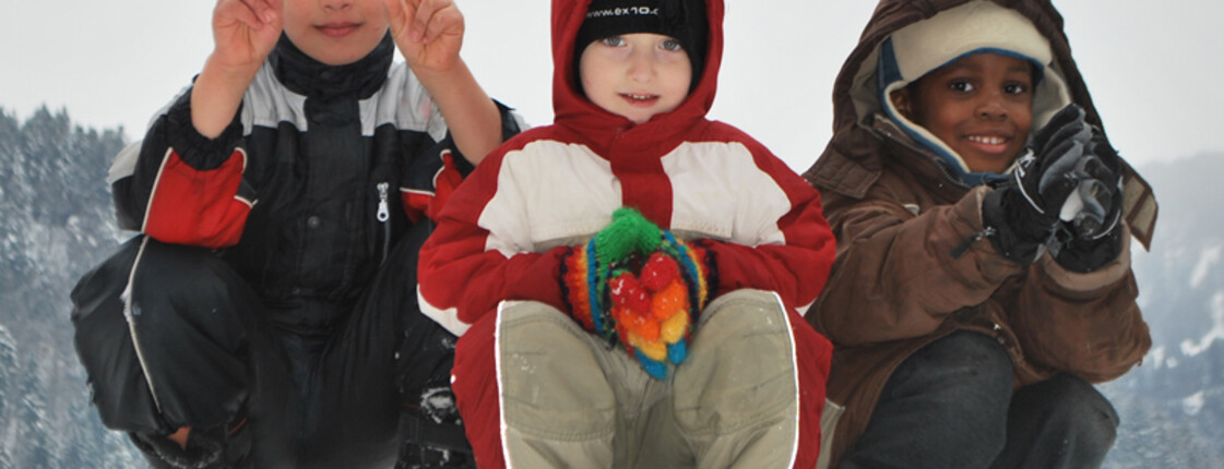 Drei Kinder unterschiedlicher Ethnien spielen im Schnee. Sie tragen dicke Winterbekleidung, die schwarz, weiß, rot und braun ist. Sie sitzen dabei in einer Hocke auf einem Schneehügel. Alle drei Kinder sind erfreut und haben Spaß. Einer der drei Jungs zeigt noch das Victory Zeichen mit beiden Händen. 