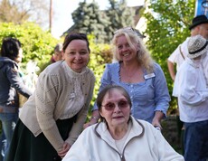 Eine Bewohnerin sitzt im Garten des Pflegewohnhauses, begleitet von der Hausleiterin und einer weiteren Mitarbeiterin, während sie gemeinsam das Maibaumfest im Freien erleben.