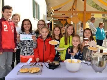 Kinder der Volksschule Göfis an einem Stand in der Gemeinde. Sie halten dabei Lebensmittel wie Marmelade und Pfannkuchen in die Kamera. 