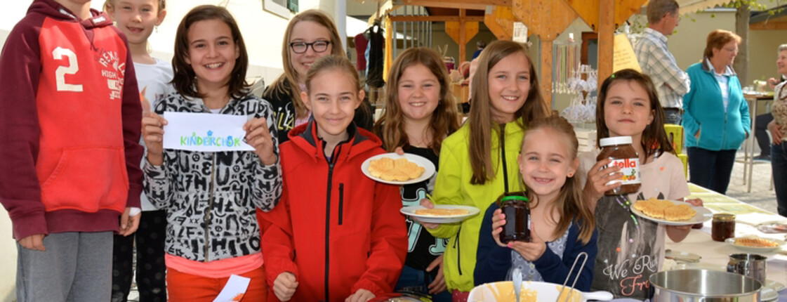 Kinder der Volksschule Göfis an einem Stand in der Gemeinde. Sie halten dabei Lebensmittel wie Marmelade und Pfannkuchen in die Kamera. 
