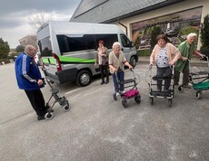 Eine Gruppe von Bewohnerinnen des Pflegewohnhauses ist gemeinsam mit Begleitpersonen auf dem Weg zum Café. Die Gruppe steht vor einem Transporter und bereitet sich mit Rollatoren auf den kurzen Fußweg vor.