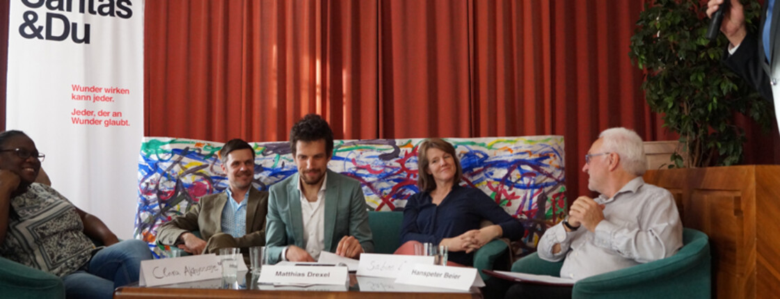 Mitarbeiter der Caritas und andere Interessensvertreter sitzen vor einem roten Vorhand um einen Tisch. Daneben ist ein Mann mit einem Mikrofon zu sehen.