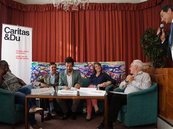 Mitarbeiter der Caritas und andere Interessensvertreter sitzen vor einem roten Vorhand um einen Tisch. Daneben ist ein Mann mit einem Mikrofon zu sehen.