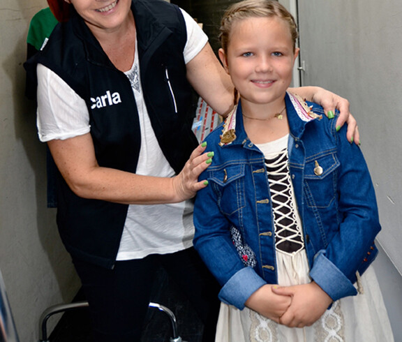 Ein kleines junges Mädchen trägt ein Kleid. Hinter ihr steht eine Frau mittleren Alters, die für Carla arbeitet.