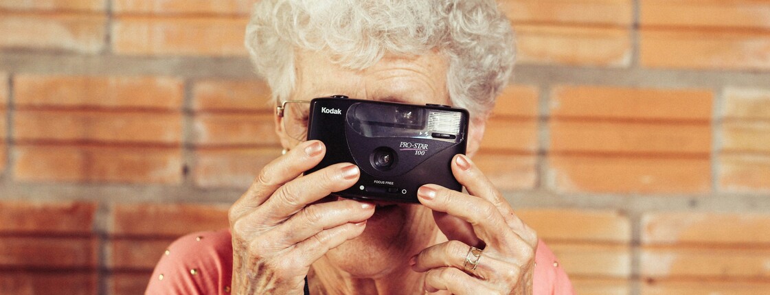 Close-up einer älteren Frau mit grauen Haaren, die durch den Sucher einer Kamera blickt und ein Foto macht.