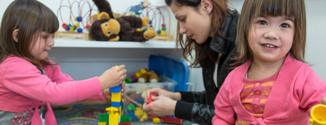 Eine junge Frau mit braunen Haaren spielt am Boden mit zwei kleinen Mädchen mit Lego. Die Frau trägt eine schwarze Lederjacke und die Mädchen rosa Oberteile. 
