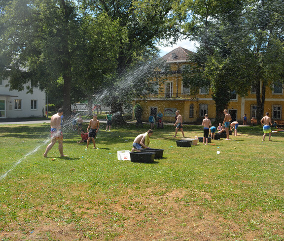Wasserfontäne auf Wiese, im Hintergrund viele Kinder in Badebekleidung