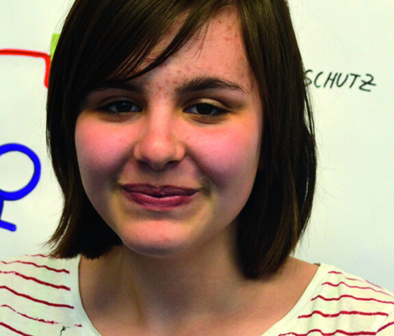 Eine Jugendliche blickt in die Kamera. Sie trägt mittellange schwarze Haare. Sie trägt ein gestreiftes Oberteil. Sie steht vor einem Flipchart. 