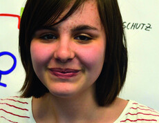 Eine Jugendliche blickt in die Kamera. Sie trägt mittellange schwarze Haare. Sie trägt ein gestreiftes Oberteil. Sie steht vor einem Flipchart. 