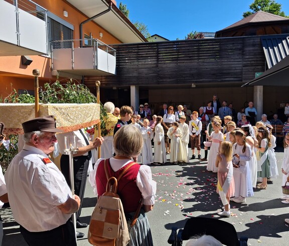Vor dem Pflegewohnhaus versammelt sich eine festliche Prozession mit Erstkommunionskindern und Gläubigen zu einer feierlichen Segnung im Freien.