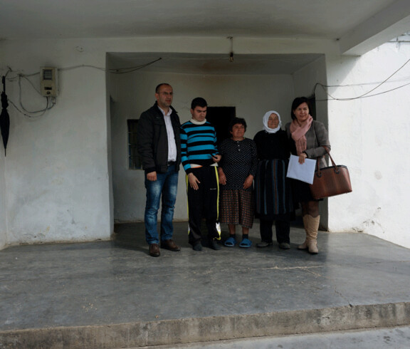 Eine Familie mit einem behinderten jungen Mann steht gemeinsam mit zwei Caritas-Mitarbeitenden vor einem schlichten Haus.