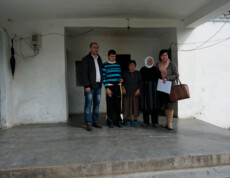 Eine Familie mit einem behinderten jungen Mann steht gemeinsam mit zwei Caritas-Mitarbeitenden vor einem schlichten Haus.