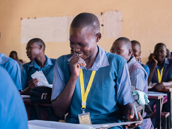 Ein junge in blauer Schuluniform grinst und wendet den Blick ab. Er sitzt in einem Klassenzimmer mit vielen anderen Kindern.