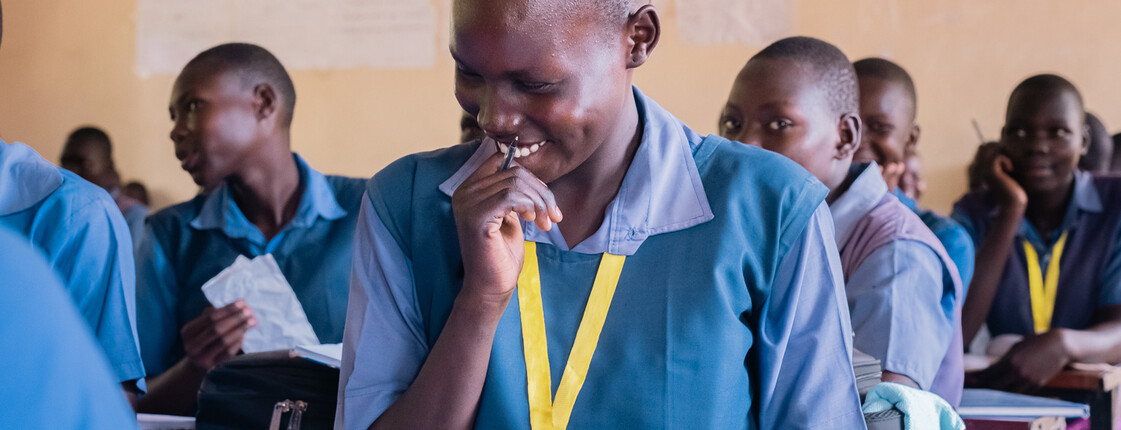 Ein junge in blauer Schuluniform grinst und wendet den Blick ab. Er sitzt in einem Klassenzimmer mit vielen anderen Kindern.