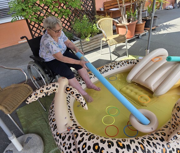 Eine Bewohnerin eines Pflegewohnhauses kühlt ihre Füße in einem Planschbecken und übt dabei spielerisch Geschicklichkeit mit verschiedenen Wasserutensilien.