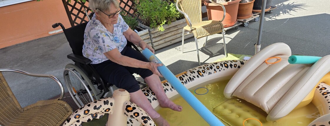 Eine Bewohnerin eines Pflegewohnhauses kühlt ihre Füße in einem Planschbecken und übt dabei spielerisch Geschicklichkeit mit verschiedenen Wasserutensilien.