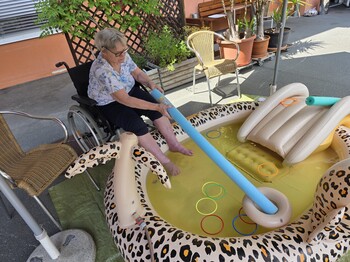 Eine Bewohnerin eines Pflegewohnhauses kühlt ihre Füße in einem Planschbecken und übt dabei spielerisch Geschicklichkeit mit verschiedenen Wasserutensilien.