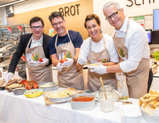 Vier Personen bereiten Speisen mit Tomaten und Brot bei Verkostung im SPAR-Supermarkt in Villach zu.