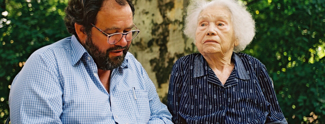 Ein Mann mittleren Alters mit einer Brille, Vollbart, schwarzen Haaren und einem blauen Hemd ließt aus einem Buch einer Dame im hohen Alter vor. Die Dame hat weiße Haare, ist dunkel gekleidet und sitzt in einem Rollstuhl. 