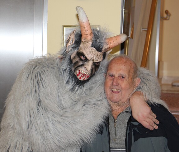 Krampus mit hellgrauem Fell steht neben einem Bewohner des Pflegewohnhauses und legt freundlich seine Hand über dessen Schulter.
