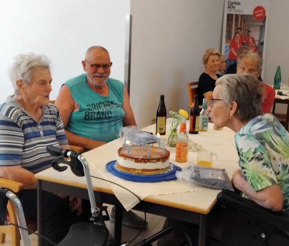 Bewohner*innen sitzen am Tisch, Geburtstagskuchen mit Kerzen wird gefeiert, Getränke stehen bereit im Pflegewohnhaus.
