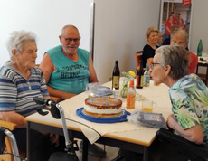 Bewohner*innen sitzen am Tisch, Geburtstagskuchen mit Kerzen wird gefeiert, Getränke stehen bereit im Pflegewohnhaus.