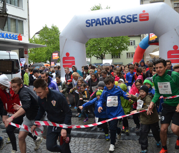Eine große Ansammlung von Jugendlichen befindet sich in einer Innenstadt. Sie alle nehmen teil an einem Laufwettbewerb. Alle haben sie Wettkampfnummern. 