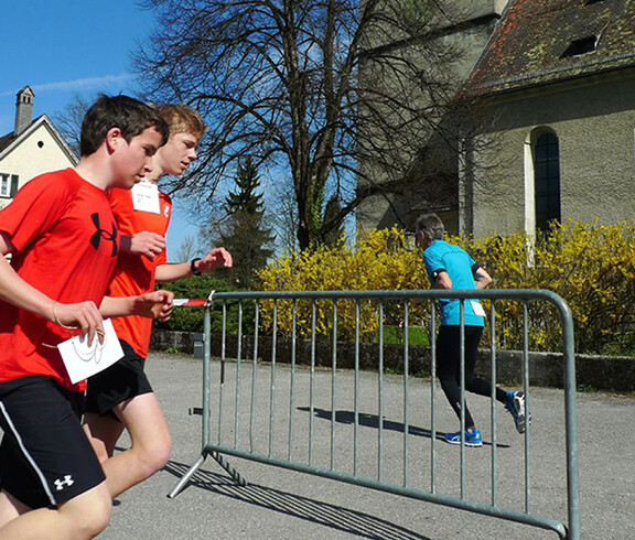 Zwei junge Burschen rennen in Sportbekleidung. Ein Junge hält Kopfhörer in seiner Hand. Gegenüber rennt ein Mann in die andere Richtung.