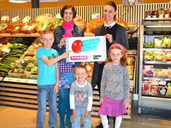 Caritas-Mitarbeiterin Doris Jenny stellt mit einer Sutterlüty-Angestellten die Aktion "Aufrunden" vor. Sie halten dabei ein Schild in der Hand, das die Aktion illustirert, Im Hintergrund befindet sich ein Regal mit Lebensmitteln. Auch kleine Kinder im Kinder- und Spielgruppenalter sind auf dem Bild und stehen bei den Erwachsenen. 