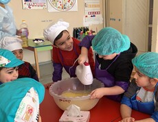 Kindergartenkinder, die verschiedene Kochmützen tragen, stehen um einen Tisch. Ein Kind rührt Teig mit einem Stabmixer.