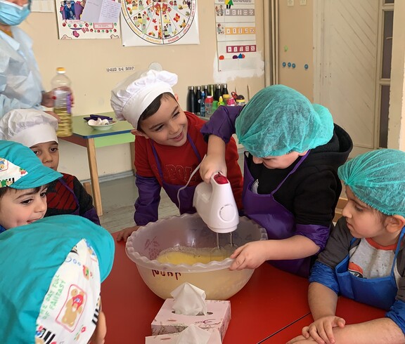 Kindergartenkinder, die verschiedene Kochmützen tragen, stehen um einen Tisch. Ein Kind rührt Teig mit einem Stabmixer.